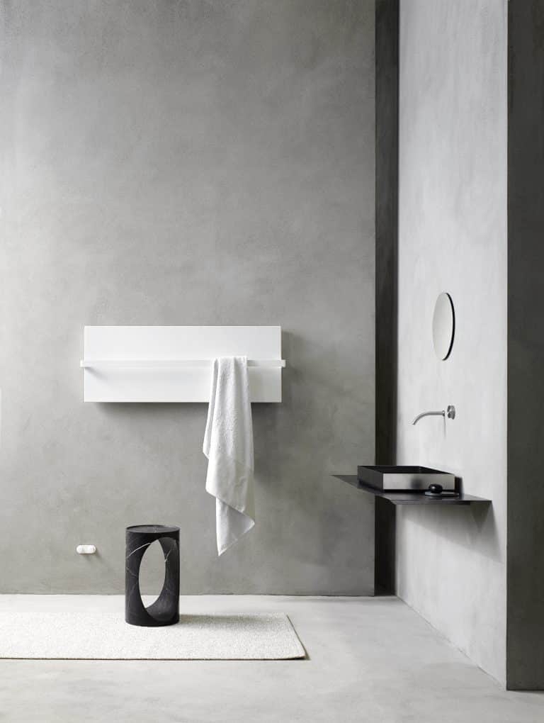 Betonová stěrka v koupelně, bílý radiátor s držákem na ručník, stříbrné umyvadlo na desce, kulaté zrcadlo