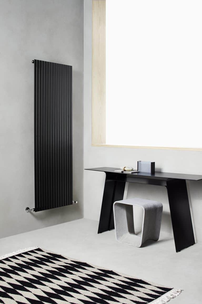 Černý žebrový radiátor na zdi, stůl se stoličkou, černobílý koberec