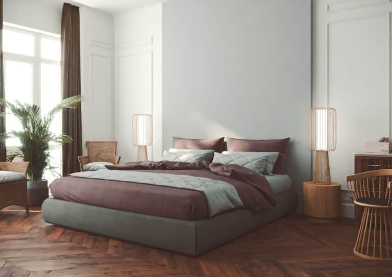 Ložnice s manželskou postelí, fialové povlečení, dřevěný nábytek a lampy, šedá stěna za postelí z umělého kamene