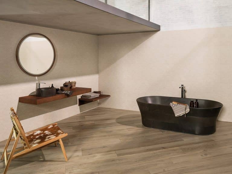 Koupelna s dlažbou s imitací dřeva, béžový obklad, černá vana do prostoru, kulaté zrcadlo, křeslo se zvířecí kůží