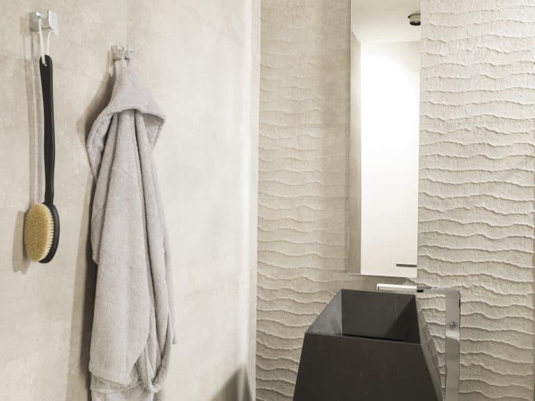 Koupelna s béžovým obkladem, černé kamenné umyvadlo, obdélníkové zrcadlo, baterie z podlahy, pověšený šedý župan a kartáč