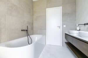 Koupelna s bílým mramorem na podlaze, bílá oválná umyvadla na desce z umělého kamene, dřevěná skříňka pod umyvadlo, rohová vana, černá vanová baterie, betonový obklad